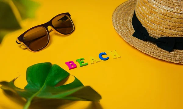 Αξεσουάρ παραλίας: ψάθινο καπέλο, γυαλιά ηλίου, η παραλία επιγραφή και ένα πλατύ φύλλο τέρας ένα φυτό για τη σκιά σε ένα φωτεινό κίτρινο φόντο σε μια ηλιόλουστη μέρα, επάνω όψη, αντίγραφο χώρου — Φωτογραφία Αρχείου