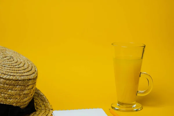 Έννοια της χαλάρωσης και της δίψας στην παραλία. ένα ποτήρι λεμονάδα σε κίτρινο φόντο με ψάθινο καπέλο και λευκό σημειωματάριο. πάνω όψη — Φωτογραφία Αρχείου