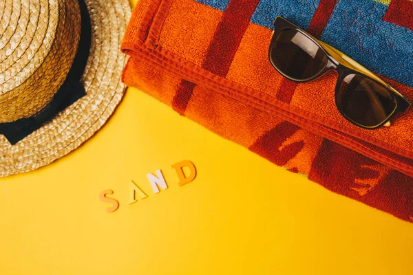 Έννοια της χαλάρωσης στην παραλία. επιγραφή άμμο σε κίτρινο φόντο με γυαλιά ηλίου, μια πετσέτα και ένα ψάθινο καπέλο. πάνω όψη — Φωτογραφία Αρχείου