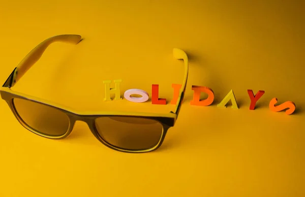 The inscription Holidays from multicolored bright wooden letters lie on a bright yellow background with sunglasses. Top view. Copy space. holiday vacation concept