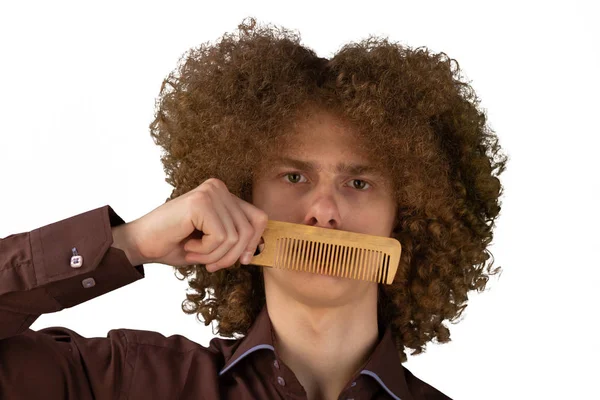 Πορτρέτο ενός σγουρά μαλλιά νεαρό άνδρα με μια ξύλινη χτένα κοντά στο στόμα του με υπέροχα μαλλιά σε ένα λευκό απομονωμένο φόντο. Ανδρική ιδέα φροντίδας μαλλιών — Φωτογραφία Αρχείου