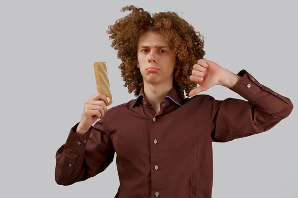 Портрет кудрявого молодого человека с деревянной расческой в великолепных волосах, недовольного результатом на сером изолированном фоне. мужской уход за волосами — стоковое фото