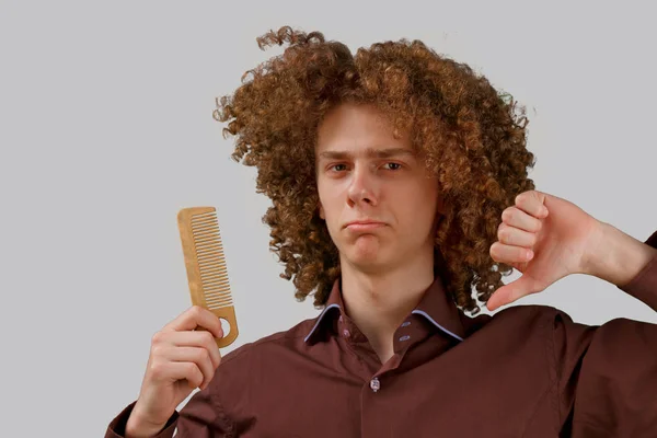 Retrato de un joven de pelo rizado con un peine de madera en magnífico pelo insatisfecho con el resultado sobre un fondo gris aislado. concepto de cuidado del cabello masculino — Foto de Stock