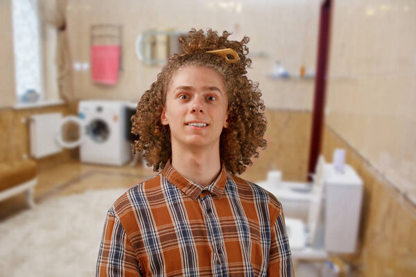 Портрет кудрявого молодого человека с деревянной расчёской в полных волосах, улыбающегося в ванной. мужской уход за волосами
