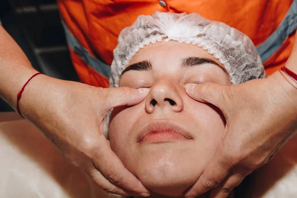 Especialista spa está fazendo massagem facial para uma jovem em um boné médico. tratamentos de beleza close-up — Fotografia de Stock