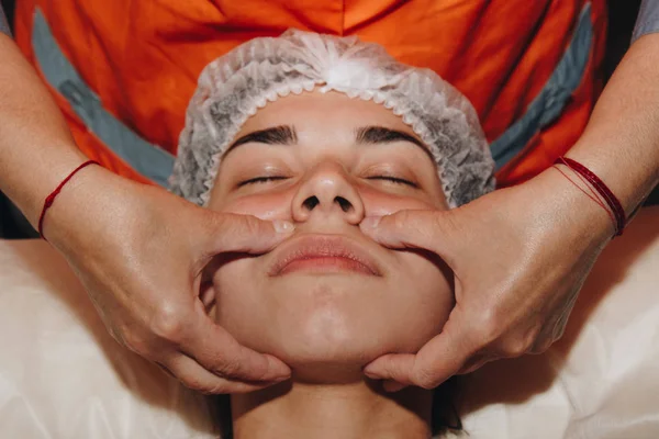 Спа-специалист делает массаж лица молодой девушке в медицинской шапке. косметические процедуры вблизи — стоковое фото
