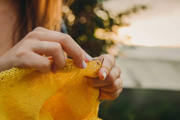 Une jeune fille aux longs cheveux blonds tricote un pull jaune dans le jardin en été. femme fait des vêtements avec les mains gros plan — Photo