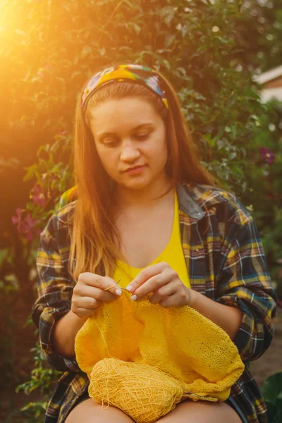 Ένα νεαρό κορίτσι με μακριά ξανθά μαλλιά πλέκει ένα κίτρινο πουλόβερ στον κήπο το καλοκαίρι. γυναίκα κάνει τα ρούχα με τα χέρια κοντινό — Φωτογραφία Αρχείου