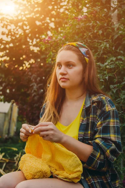 Ένα νεαρό κορίτσι με μακριά ξανθά μαλλιά πλέκει ένα κίτρινο πουλόβερ στον κήπο το καλοκαίρι. γυναίκα κάνει τα ρούχα με τα χέρια κοντινό — Φωτογραφία Αρχείου
