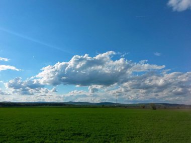 Tarım arazisi yeşil ekinlerle kaplı, güneşli bir günde, arka planda verici direkleri, ormanlar ve dağlar, görüntü