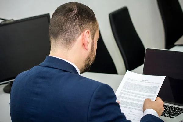 businessman with a beard in the office reads a contract before signing on a laptop background