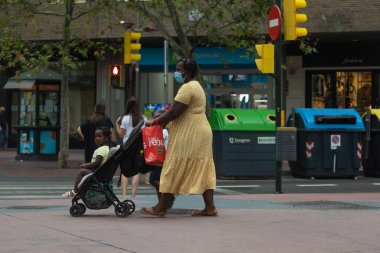 Zaragoza, İspanya - 18 Ağustos 2020: Coronavirus salgını nedeniyle yüzü maskeli siyah bir göçmen kadın, çocuğuyla birlikte Zaragoza 'nın orta bölgesine bir bebek arabasını itti..