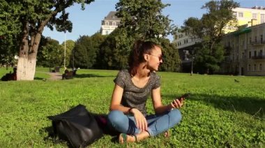 Güzel gülümseyen genç kız parkta çim üzerinde oturur ve onun favori melodi telefonda dinler.