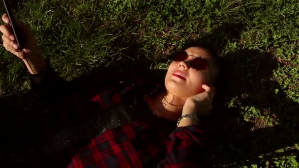 Belle jeune fille en lunettes de soleil couchée sur l'herbe et écoutant de la musique sur son téléphone . — Video