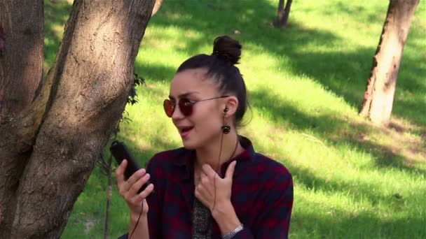 Jeune belle fille écoutant de la musique dans son smartphone, chante et danse tout en étant assis sur un banc de parc . — Video