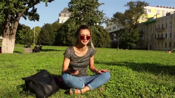 Krásná mladá dívka, která nosí sluneční brýle sedí na trávě v parku, na její oblíbené melodie v telefonu. — Stock video