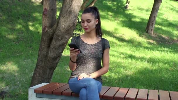 Όμορφη κοπέλα κάθεται σε ένα παγκάκι στο πάρκο και την περιήγηση ταινία κοινωνικής δικτύωσης στο τηλέφωνό της. — Αρχείο Βίντεο