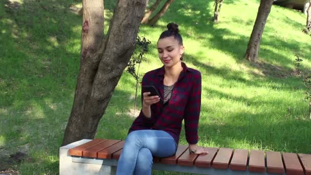 Menina bonita nova em uma camisa xadrez sorrindo e navegando na fita de redes sociais em seu smartphone . — Vídeo de Stock
