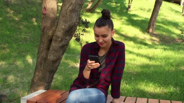Menina bonita nova em uma camisa xadrez senta-se em um banco no parque, ativamente reescrito no bate-papo na internet — Vídeo de Stock