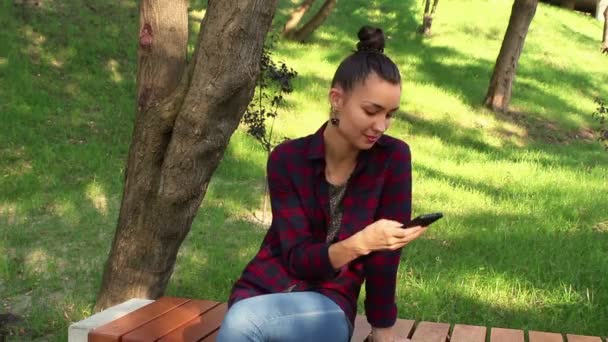 Jeune belle fille dans une chemise à carreaux souriant et naviguant sur la bande des réseaux sociaux dans son smartphone . — Video
