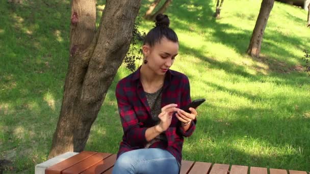 Joven chica hermosa en una camisa a cuadros está sentado en un banco del parque, sonriendo y escribiendo un mensaje en su teléfono inteligente . — Vídeos de Stock