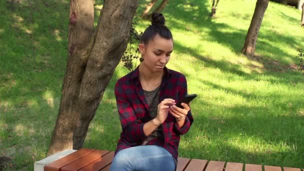 Krásná mladá dívka v kostkované košili sedí na lavičce v parku, aktivně přepsán v internetové konverzace — Stock video