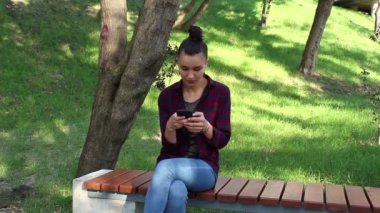 Genç güzel kız ekose gömlekli aktif Internet sohbet yeniden parkta bir bankta oturur.
