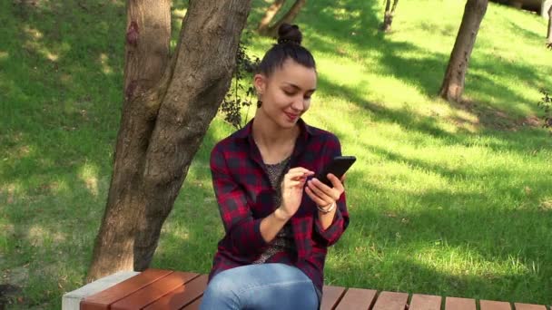 Young beautiful girl is sitting on a bench in the park and browsing the social networking tape in her phone. — Stock Video