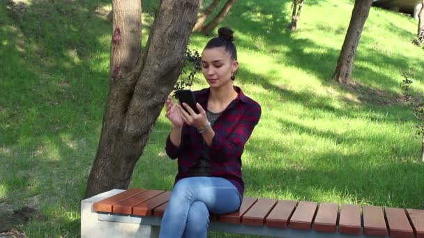 Menina bonita nova em uma camisa xadrez sorrindo e navegando na fita de redes sociais em seu smartphone . — Vídeo de Stock
