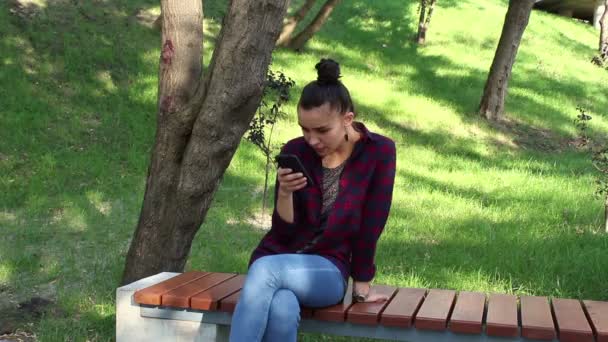 Ekose gömlekli genç güzel kız gülümseyerek ve onun smartphone içinde mesaj yazarken bir bankta oturuyor. — Stok video