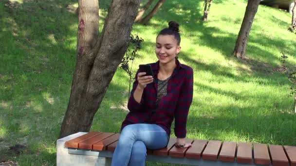 Krásná mladá dívka v kostkované košili sedí na lavičce v parku, usmívající se a aktivně přepsaný v internetové konverzace. — Stock video