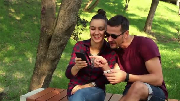Para zakochanych pokazać zdjęcia na ich smartfonach z sobą, siedząc na ławce w parku. — Wideo stockowe