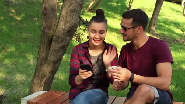 Casal apaixonado mostrar fotos em seus smartphones uns com os outros, sentado em um banco de parque . — Vídeo de Stock