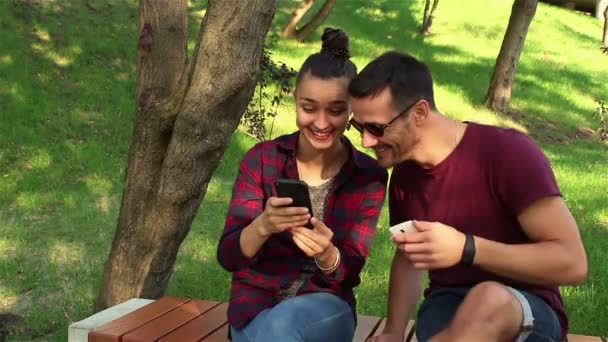 Pareja enamorada mostrar fotos en sus teléfonos inteligentes entre sí, sentado en un banco del parque . — Vídeos de Stock