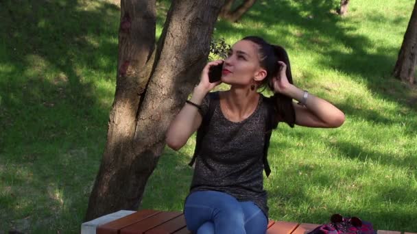 Giovane ragazza felice che parla al telefono, sorride e si tocca i capelli, seduta su una panchina del parco . — Video Stock