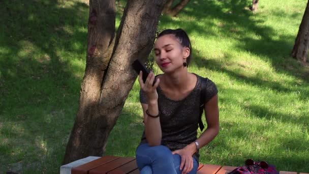 年轻美丽的女孩在电话里说话 微笑着触摸她的头发 坐在公园的长椅上 — 图库视频影像