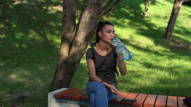 Mooi meisje is drinkwater zittend op een bankje in het park. — Stockvideo
