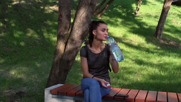 Jeune belle fille boit de l'eau assise sur un banc dans le parc . — Video