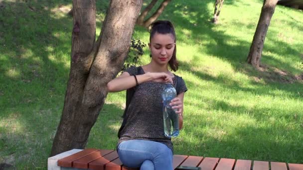 Mooi meisje is drinkwater zittend op een bankje in het park. — Stockvideo