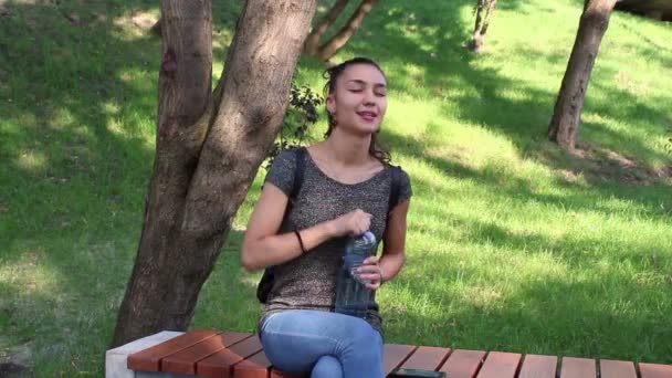 Jeune belle fille secoue une bouteille en plastique avec de l'eau, assise sur un banc à l'ombre d'un arbre . — Video