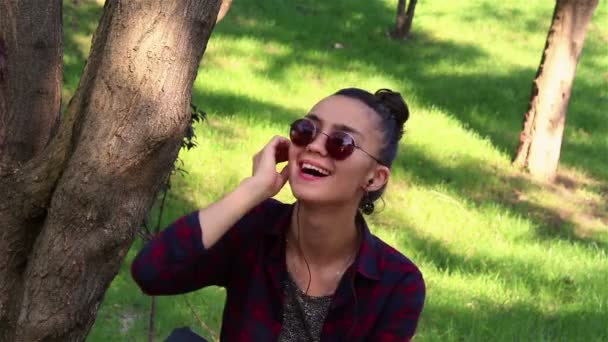Hermosa chica escuchando música a través de auriculares, bailando y cantando mientras está sentada en un banco del parque . — Vídeo de stock