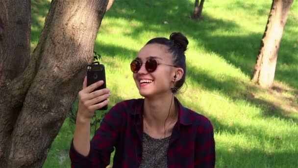 戴着太阳镜的美丽女孩, 积极地笑着, 透过社交网络上的新闻, 坐在公园的长椅上. — 图库视频影像
