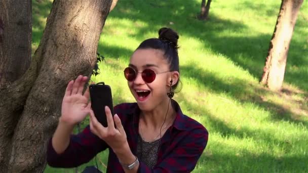 Hermosa chica en gafas de sol, riendo activamente, mirando a través de las noticias en las redes sociales, sentado en un banco del parque . — Vídeo de stock