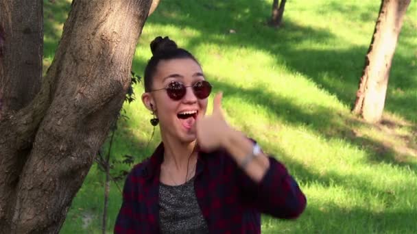 Hermosa chica con gafas de sol, riendo activamente, y mostrando el pulgar hacia arriba mientras está sentado en un banco en el parque — Vídeo de stock