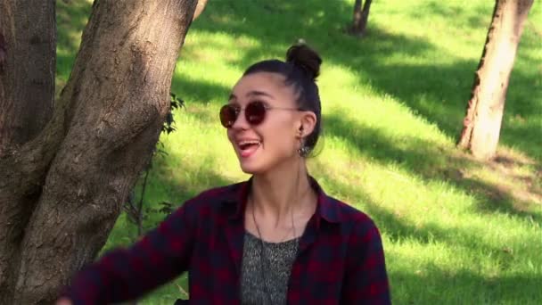 Belle fille en lunettes de soleil, riant activement, et montrant pouce levé tout en étant assis sur un banc dans le parc — Video