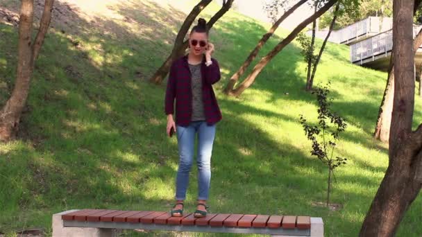 Chica joven bailando de pie en un banco en el parque, escuchando música en su teléfono inteligente . — Vídeos de Stock