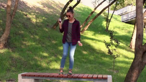 Chica joven bailando de pie en un banco en el parque, escuchando música en su teléfono inteligente . — Vídeos de Stock