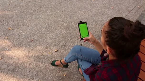 Young beautiful girl listening to music on her smartphone, sitting on a bench in the park. — Stock Video