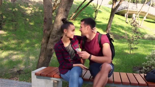 Jóvenes amantes besándose sentados en un banco del parque . — Vídeos de Stock