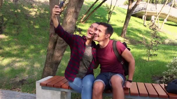 Jóvenes amantes hacen una foto sentados en un banco del parque . — Vídeos de Stock